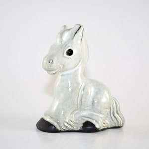 May include: A ceramic figurine of a resting donkey. The donkey is light gray with black hooves and dark eyes. The figurine is a decorative item, likely for a shelf or display.