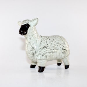 May include: A ceramic sheep figurine with a white body and black face, ears, and hooves. The sheep has a textured, wool-like appearance. The figurine is standing on a white surface.