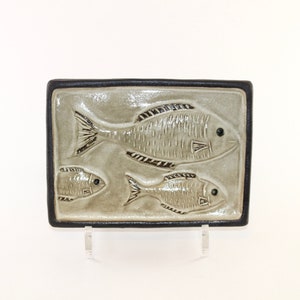 May include: A rectangular ceramic tile featuring a relief design of three fish. The fish are in varying sizes, with the largest at the top. The tile has a gray background with black outlines and a black border.