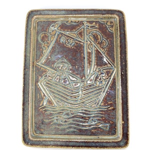 Könnte beinhalten: Rechteckige Keramikschale mit einem erhabenen Reliefdesign eines Segelbootes mit einem Seemann. Die Schale ist braun mit einem helleren, blau-grauen Design. Der Text "R 57" ist auf dem Segel sichtbar. Die Schale hat einen erhabenen Rand.