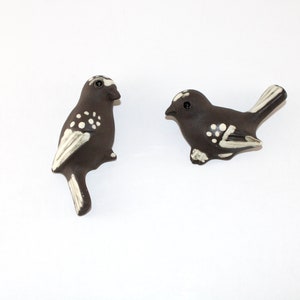 May include: Two ceramic bird figurines. The birds are dark brown with white accents on the wings, tail, and body. One bird is perched upright, while the other is in a resting position. The background is plain white.
