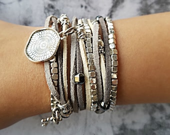 Puede incluir: Un brazalete de múltiples capas con un dije de plata y cuentas. El brazalete está hecho de cordón de cuero gris y blanco y cuentas de plata.