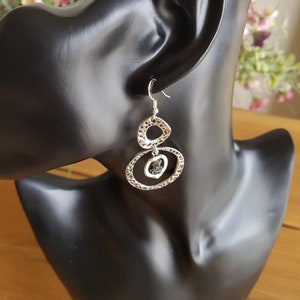 May include: Silver dangle earrings with a textured finish. The earrings feature two connected circles with a black gemstone in the center.