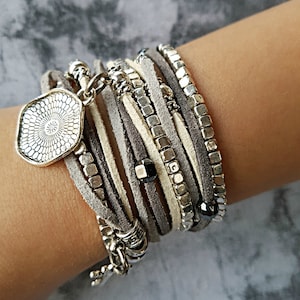 Op de afbeelding: Een meerlaags armband met grijze en witte leren koorden, zilveren kralen en een zilveren bedel met een bloemenmotief.