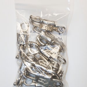 May include: A bag of silver-toned metal clasps with a spring-loaded gate. The clasps are designed to be used for attaching straps or leashes to items.