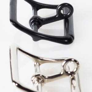 Puede incluir: Dos hebillas de cinturón de metal, una negra y otra plateada. Ambas hebillas tienen un diseño similar con una forma curva y un pequeño bucle en el lateral.
