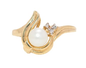 May include: A gold-toned ring featuring a large, round, white pearl as the focal point. The ring has a flowing, asymmetrical design with a cluster of small, clear gemstones on one side.