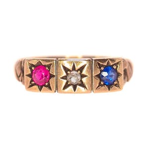 May include: A gold ring featuring three square settings. The settings hold a pink gemstone, a clear gemstone, and a blue gemstone, each surrounded by a starburst design. The ring has a textured band.