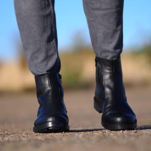 May include: A pair of black leather ankle boots with a low heel and a side zipper. The boots are worn with gray pants. The image is taken outdoors with a blurred background.