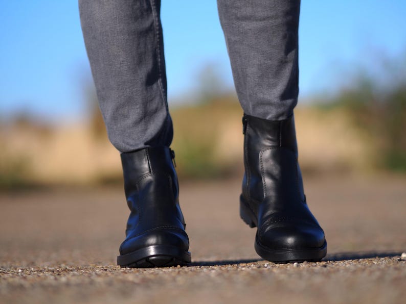 May include: A pair of black leather ankle boots with a low heel and a side zipper. The boots are worn with gray pants. The image is taken outdoors with a blurred background.