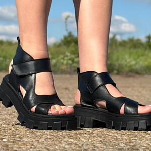 Pu&ograve; includere: Sandali platform in pelle nera con suola carrarmato. I sandali hanno un cinturino alla caviglia e un design ritagliato. L'immagine &egrave; scattata all'aperto in una giornata di sole.