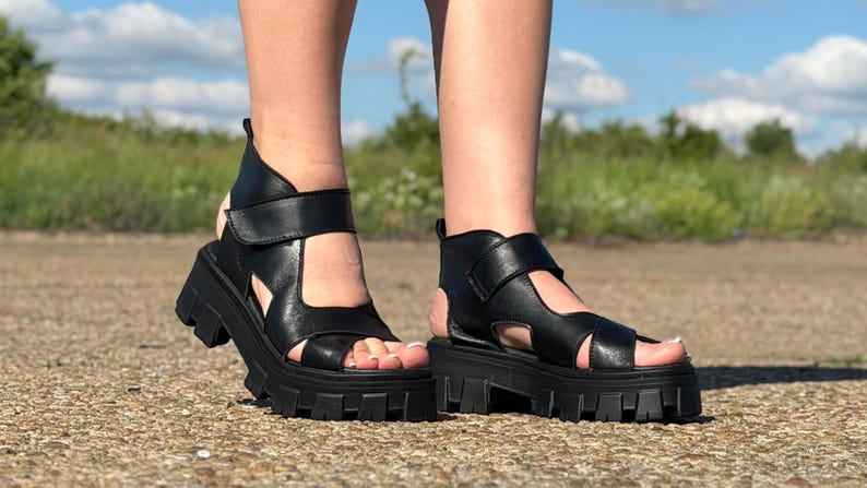 Pu&ograve; includere: Sandali platform in pelle nera con suola carrarmato. I sandali hanno un cinturino alla caviglia e un design ritagliato. L'immagine &egrave; scattata all'aperto in una giornata di sole.