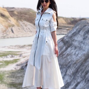 May include: A white maxi dress with a zipper down the front, a drawstring waist, and cut-out shoulders. The dress has pockets on the sides and a black trim around the collar and zipper. The model is wearing white sneakers.
