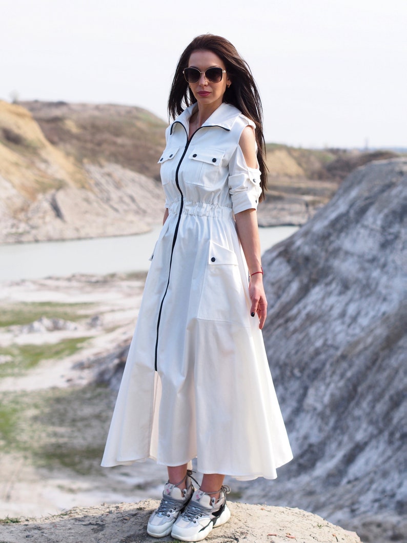 May include: A white maxi dress with a zipper down the front, a drawstring waist, and cut-out shoulders. The dress has pockets on the sides and a black trim around the collar and zipper. The model is wearing white sneakers.