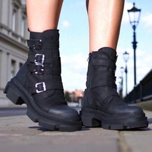 May include: Black leather ankle boots with multiple buckles and a chunky sole. The boots have a side zipper and are worn outdoors on a paved surface. The boots are a stylish and edgy footwear choice.
