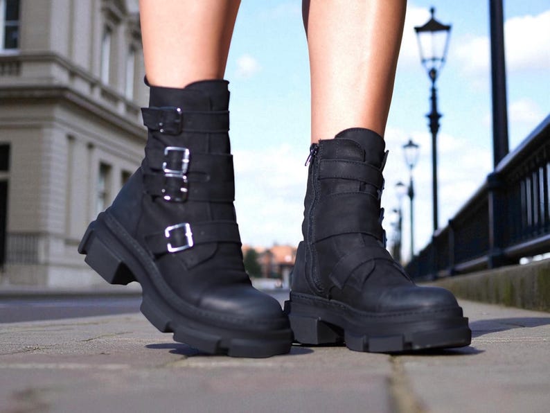 May include: Black leather ankle boots with multiple buckles and a chunky sole. The boots have a side zipper and are worn outdoors on a paved surface. The boots are a stylish and edgy footwear choice.