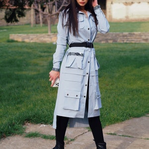May include: A light blue long-sleeved shirt dress with a black belt and black boots. The dress has multiple pockets and a button-up front. The model is wearing sunglasses.