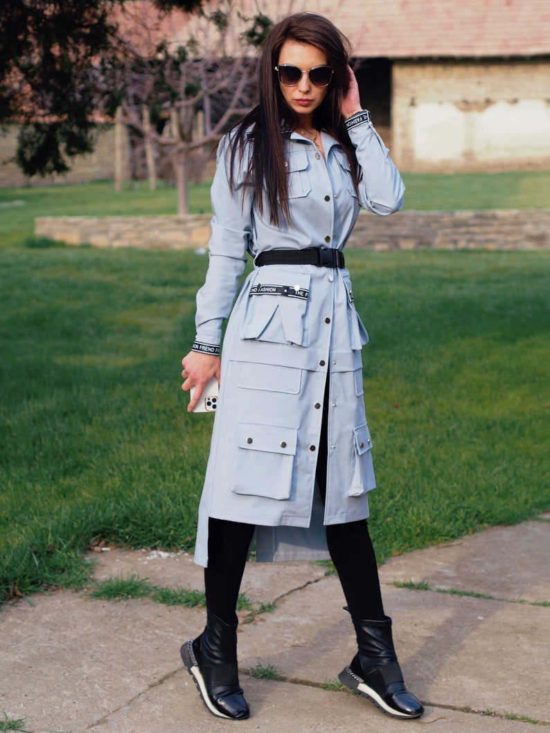 May include: A light blue long-sleeved shirt dress with a black belt and black boots. The dress has multiple pockets and a button-up front. The model is wearing sunglasses.