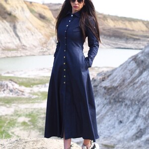 May include: A long, dark blue denim dress with a button-up front and long sleeves. The dress has a fitted bodice and a flowing skirt. The model is wearing white sneakers.