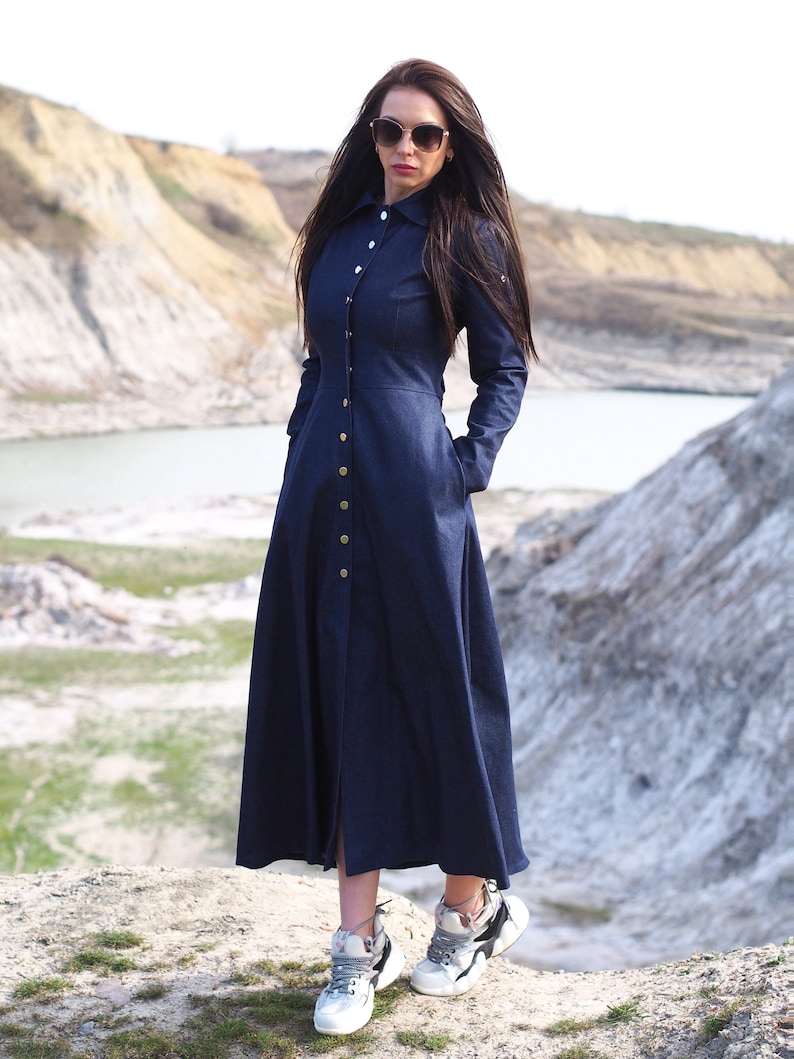 May include: A long, dark blue denim dress with a button-up front and long sleeves. The dress has a fitted bodice and a flowing skirt. The model is wearing white sneakers.