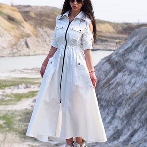 May include: A white maxi dress with a zipper down the front and a drawstring waist. The dress has a collar and short sleeves with cutouts at the shoulders. The model is wearing white sneakers.