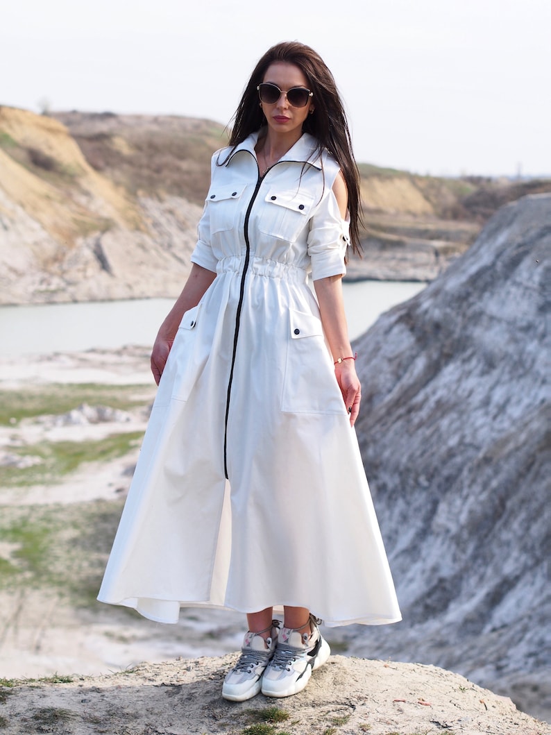 May include: A white maxi dress with a zipper down the front and a drawstring waist. The dress has a collar and short sleeves with cutouts at the shoulders. The model is wearing white sneakers.