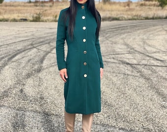 May include: A dark green long-sleeved coat featuring a high collar, a row of gold-toned buttons down the front, and two side pockets. The coat is paired with a matching dark green hat, oversized black sunglasses, and beige knee-high boots.