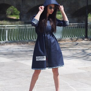 May include: A blue denim dress with a hood and a zipper closure. The dress has a drawstring waist and a white graphic print on the front that says "It's suit yourself in the world's wardrobe". The dress also has a white print along the bottom hem that says "Adventure begins your vibe vibe attracts your tribe".