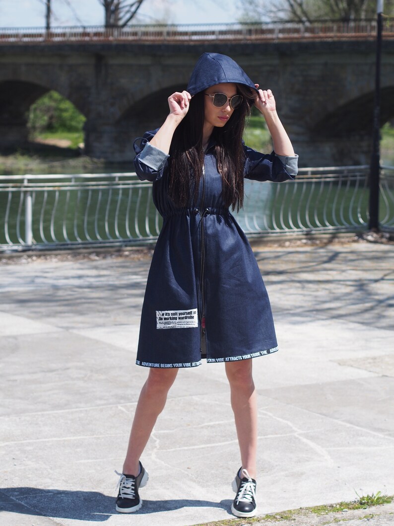 May include: A blue denim dress with a hood and a zipper closure. The dress has a drawstring waist and a white graphic print on the front that says "It's suit yourself in the world's wardrobe". The dress also has a white print along the bottom hem that says "Adventure begins your vibe vibe attracts your tribe".