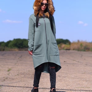 May include: A long, sage green hooded sweatshirt with a faux fur hood and a zipper closure. The sweatshirt has a logo on the left chest and a high-low hemline.