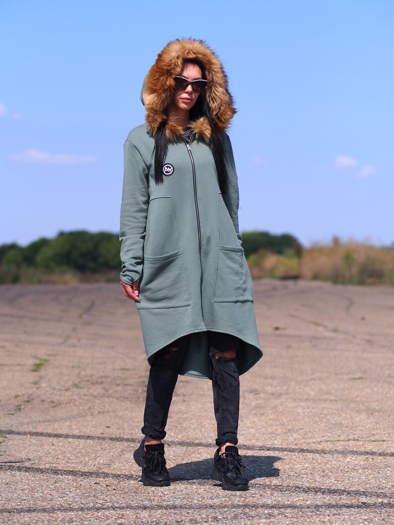 May include: A long, sage green hooded sweatshirt with a faux fur hood and a zipper closure. The sweatshirt has a logo on the left chest and a high-low hemline.