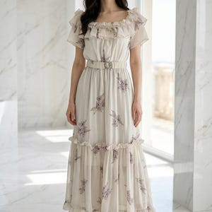 May include: A cream-colored midi dress featuring a ruffled neckline, short sleeves, and a matching fabric belt with a circular buckle. The garment is adorned with a delicate purple floral print and includes a tiered ruffled hemline.