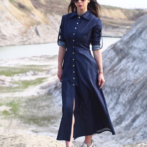 May include: A woman wearing a dark blue denim dress with a button-up front and long sleeves. She is wearing white sneakers and sunglasses. The dress has a slit up the front and is a maxi length.