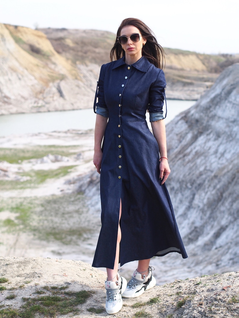 May include: A woman wearing a dark blue denim dress with a button-up front and long sleeves. She is wearing white sneakers and sunglasses. The dress has a slit up the front and is a maxi length.