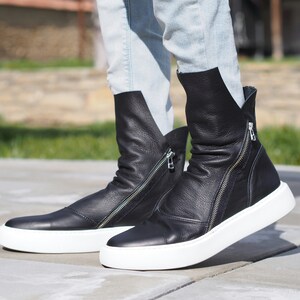 Puede incluir: Botas de tobillo de cuero negro con suelas blancas y cremalleras laterales. Las botas est&aacute;n ligeramente ca&iacute;das y tienen un aspecto informal y relajado.