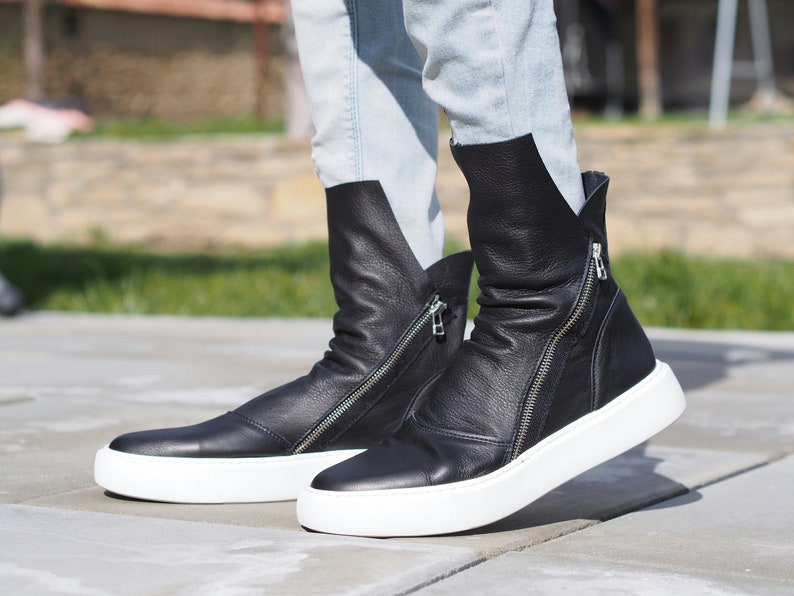 Puede incluir: Botas de tobillo de cuero negro con suelas blancas y cremalleras laterales. Las botas est&aacute;n ligeramente ca&iacute;das y tienen un aspecto informal y relajado.