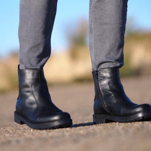 May include: Black leather ankle boots with a side zipper and a rounded toe. The boots have a thick, black sole and are worn with gray denim jeans. The boots are a classic style, suitable for various occasions.