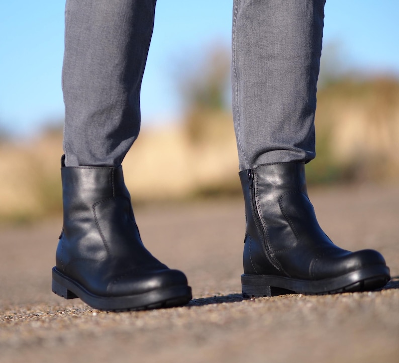 May include: Black leather ankle boots with a side zipper and a rounded toe. The boots have a thick, black sole and are worn with gray denim jeans. The boots are a classic style, suitable for various occasions.