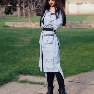 May include: A light blue long-sleeved jacket with a black belt and a black and white text detail that reads "ON TREND FASHION". The jacket has a large pocket on the front and a slit on the side. The model is wearing black leggings and black boots.
