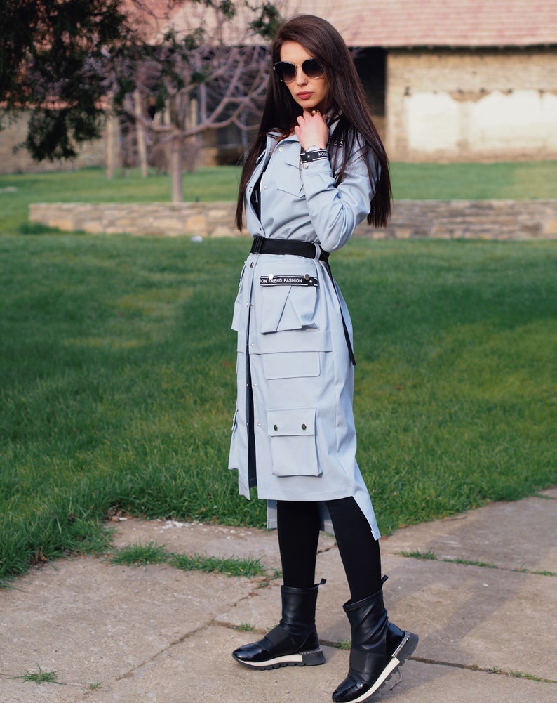 May include: A light blue long-sleeved jacket with a black belt and a black and white text detail that reads "ON TREND FASHION". The jacket has a large pocket on the front and a slit on the side. The model is wearing black leggings and black boots.