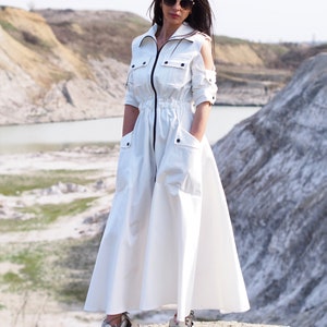 May include: A white long-sleeved dress with a zipper down the front and a drawstring waist. The dress has cut-out shoulders and large pockets on the sides. The model is wearing the dress with white sneakers.