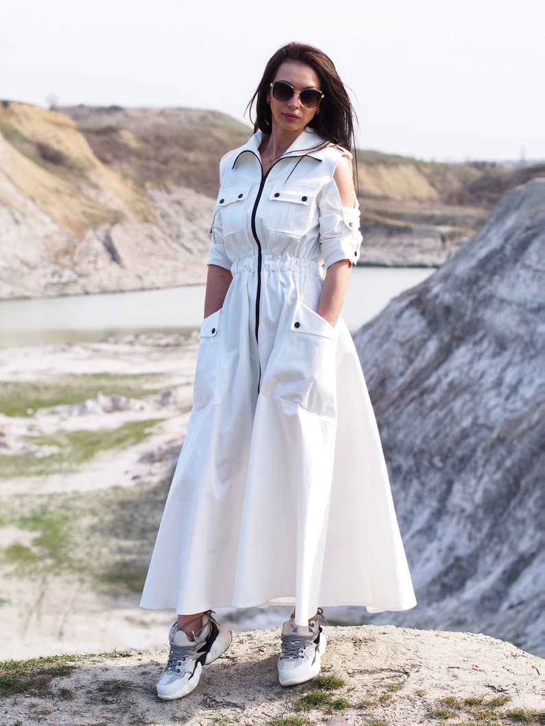May include: A white long-sleeved dress with a zipper down the front and a drawstring waist. The dress has cut-out shoulders and large pockets on the sides. The model is wearing the dress with white sneakers.