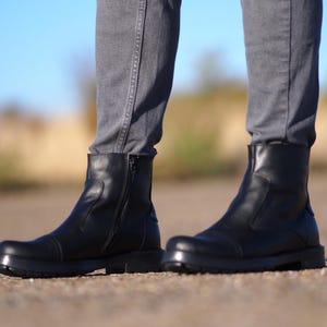 May include: Black leather ankle boots with a side zipper and a rounded toe. The boots have a thick sole and are worn with gray jeans. The boots are a classic style, suitable for various occasions.