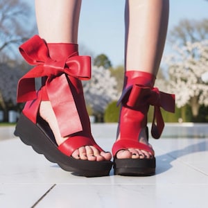 Könnte beinhalten: Rote Plateausandalen mit großen Schleifenakzenten. Die Sandalen haben eine schwarze Sohle und einen Knöchelriemen. Im Hintergrund ein weißes Gebäude und blühende Bäume.