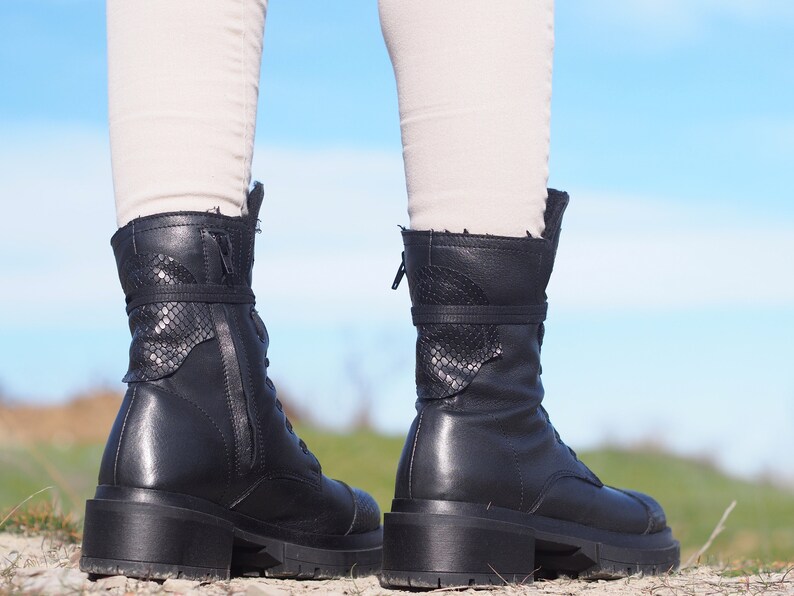 K&ouml;nnte beinhalten: Ein Paar schwarze Lederstiefel mit Rei&szlig;verschluss und Schn&uuml;rsenkeln. Die Stiefel haben ein Schlangenhautmuster an der R&uuml;ckseite des Kn&ouml;chels.