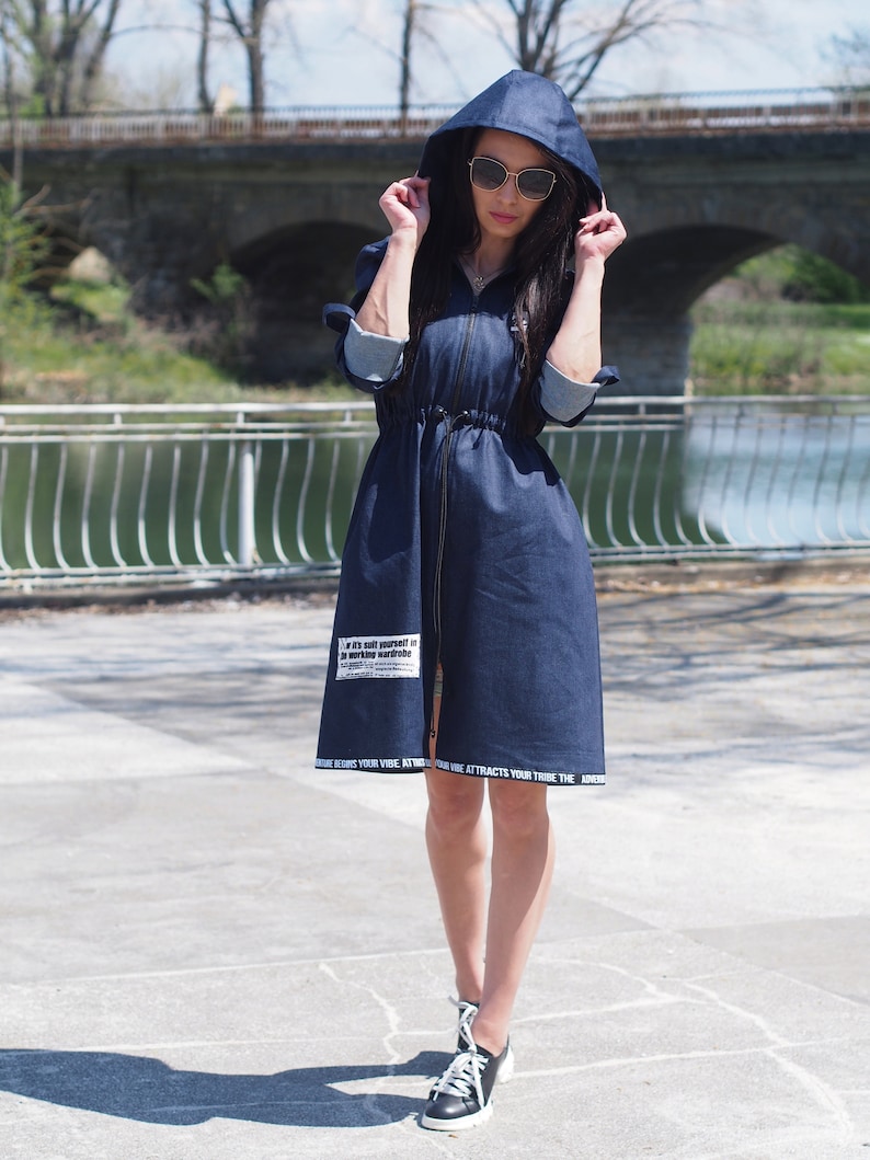 May include: A dark blue denim jacket with a hood and a zipper closure. The jacket has a drawstring waist and white text on the bottom hem and a patch on the front that says "If it's not yourself in your working wardrobe".