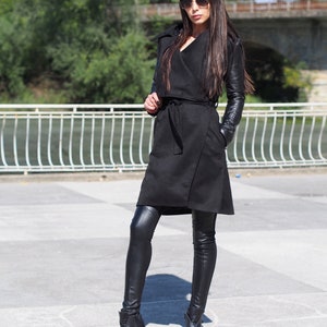 May include: A black long-sleeved coat with a wrap style and a belt. The coat has leather sleeves and is worn with black leggings and black ankle boots.