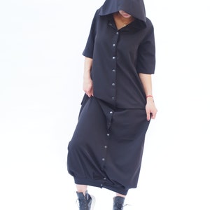Puede incluir: Un vestido negro con capucha, botones en la parte delantera y mangas largas. El vestido tiene un corte holgado y llega hasta los tobillos. La modelo lleva botas negras de tobillo.