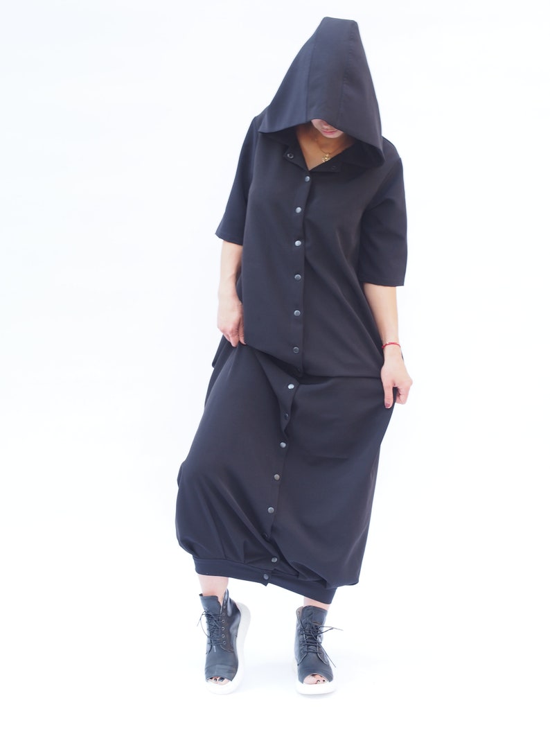 Puede incluir: Un vestido negro con capucha, botones en la parte delantera y mangas largas. El vestido tiene un corte holgado y llega hasta los tobillos. La modelo lleva botas negras de tobillo.