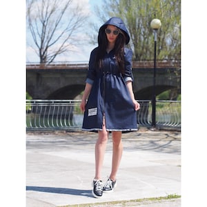 May include: A dark blue denim dress with a hood and a zipper down the front. The dress has a drawstring waist and white text along the bottom hem. The model is wearing black sneakers.